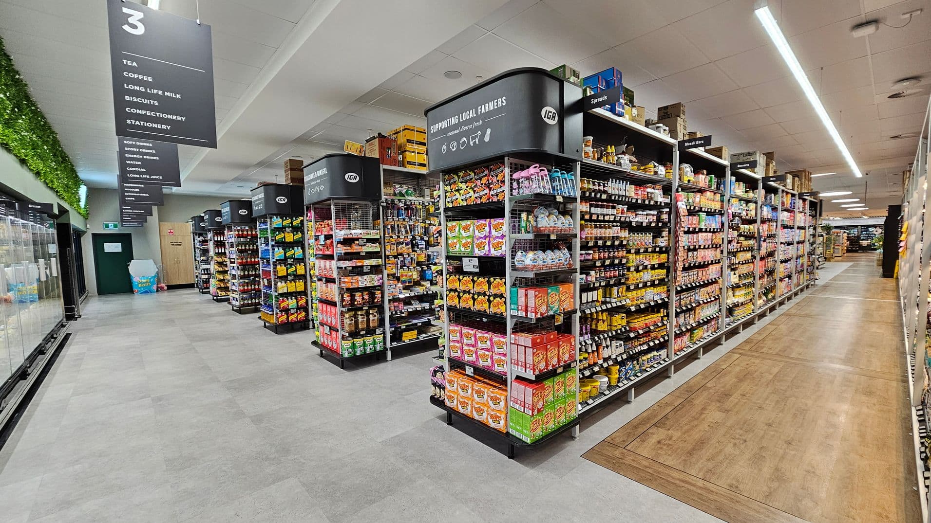 A supermarket aisle with shelves on both sides filled with various packaged goods. On the left, there are refrigerated sections with glass doors. Overhead signs indicate different product categories like tea, coffee, and confectionery. The floor is a mix of grey tiles and wood-like panels. The ceiling is fitted with fluorescent lights, and there is greenery along the top of the left wall.