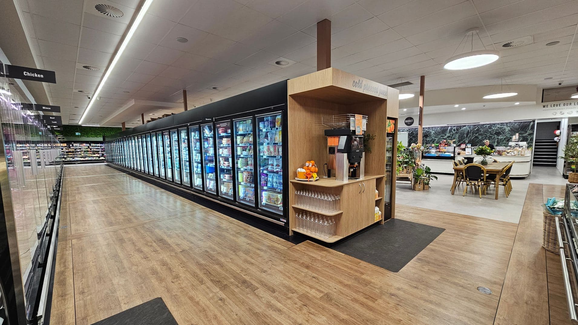 A supermarket interior with a long aisle of refrigerated display cases on the left, filled with various frozen products. Above, signs indicate sections like "Chicken" and "Mince." In the centre, a wooden kiosk offers cold pressed juice with a juicer, glasses, and bottles. On the right, a seating area with tables and chairs is visible, along with a counter and decorative plants. The floor is a mix of wood and tile, and the ceiling has circular lights and ventilation panels.