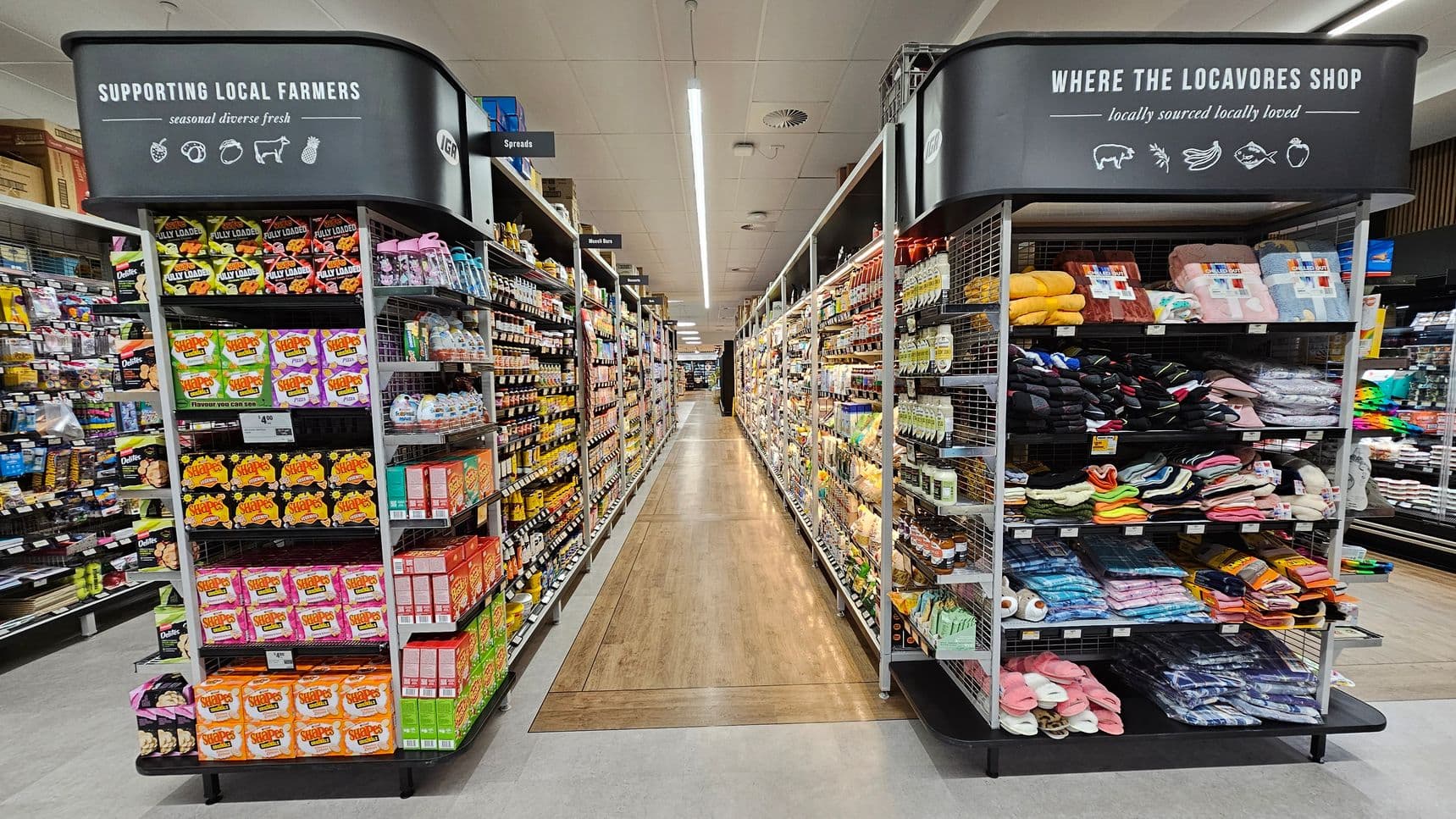 A supermarket aisle with shelves on both sides. On the left, shelves are stocked with snacks, cereals, and various packaged goods. A sign above reads "SUPPORTING LOCAL FARMERS." On the right, shelves display clothing items like socks and slippers, with a sign above saying "WHERE THE LOCAVORES SHOP." The floor is a mix of light and dark wood, and the ceiling has fluorescent lighting. The aisle extends into the distance with more products visible.