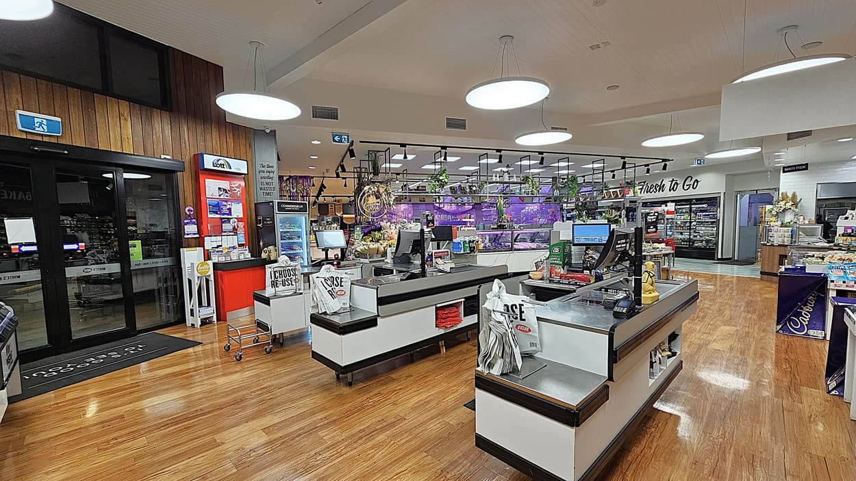 A supermarket with polished wooden floors. On the left, there is a glass entrance door, a red ATM, and a small display of magazines and brochures. In the middle, there are two checkout counters with black and white designs, each with a conveyor belt and shopping bags. The ceiling has white panels with large circular lights. On the right, there are shelves with flowers, a "Flowers To Go" sign, and a display of Cadbury Dairy Milk chocolates.