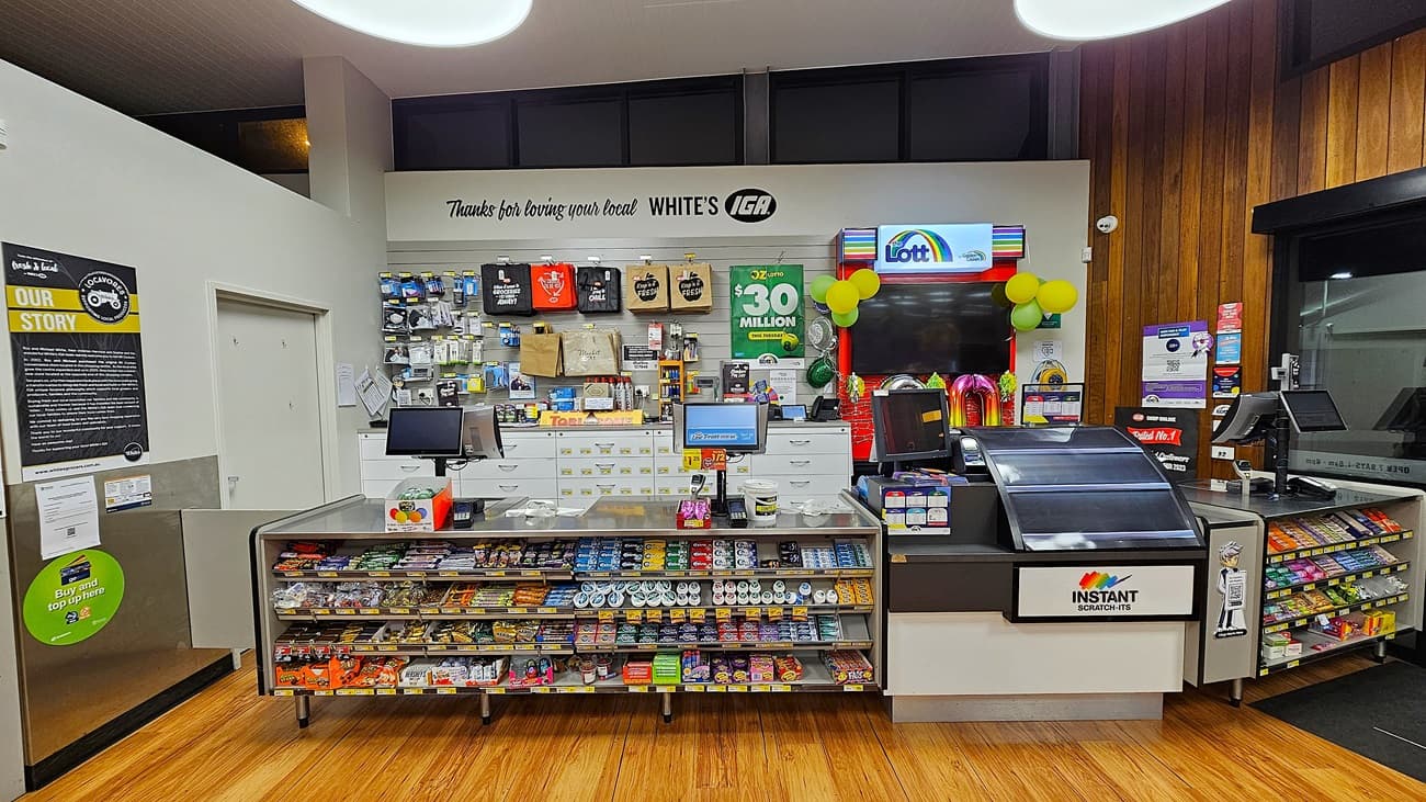 A supermarket with polished wooden floors. On the left, there is a white wall with a bulletin board and a door. In the middle, there are two checkout counters with various snacks, a display of gift cards, and a sign reading "Thanks for loving your local WHITE'S IGA." The ceiling has white panels with large circular lights. On the right, there is a lottery ticket machine with a colourful display and a window with a view outside. The background features more shelves with snacks and a "Lott" sign.