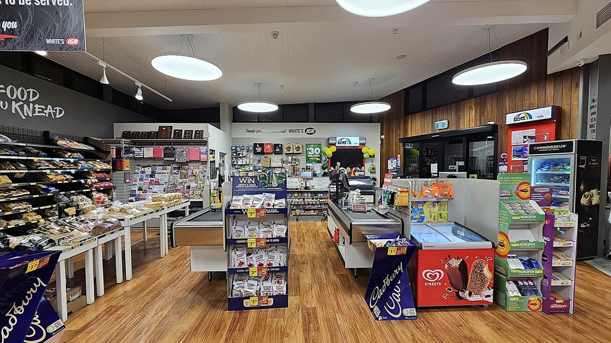 A supermarket with polished wooden floors. On the left, there are shelves with baked goods and a black wall with the words "FOOD YOU KNEAD." In the middle, there are two checkout counters with various snacks, a display of gift cards, and a sign reading "Thanks for loving your local WHITE'S IGA." The ceiling has white panels with large circular lights. On the right, there is a lottery ticket machine, a red ice cream freezer, and a window with a view outside.