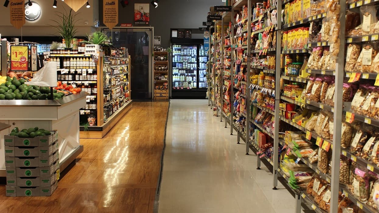 This section of the grocery store is where you will find cold drinks and snacks. There are also fruits and vegetables with signage on the wall stating, “Fresh & Local”. A rectangular stand in the middle of this section offers different fruits such as grapes, pineapples, apples, oranges, watermelons and more. Plants placed in a wooden plant pot can be seen at the sides of the fruit section. Display of chilled and frozen goods can be found on the left side of this section.