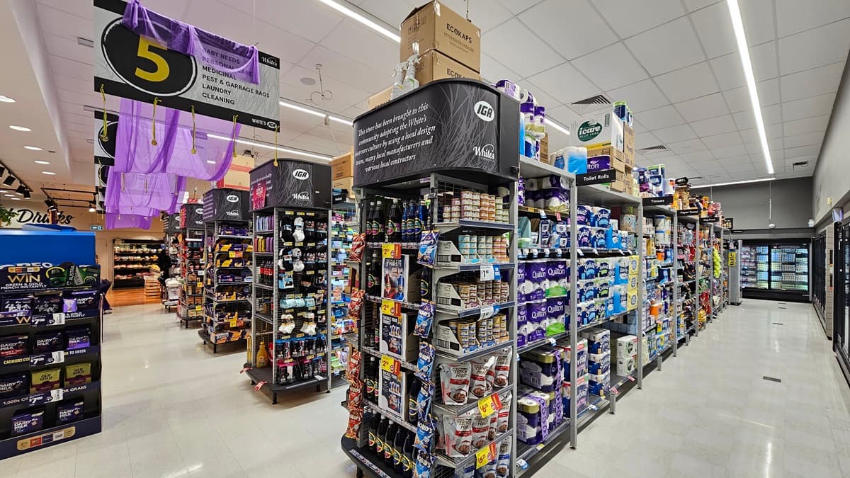 A supermarket aisle with a white tiled floor. On the left, there are shelves stocked with cleaning products and a large black sign with a yellow number 5. In the middle, there are displays of various household items, including light bulbs and batteries. The ceiling is white with rectangular fluorescent lights. On the right, there are shelves filled with toilet paper and paper towels. The background features more aisles and a refrigerated section with glass doors.