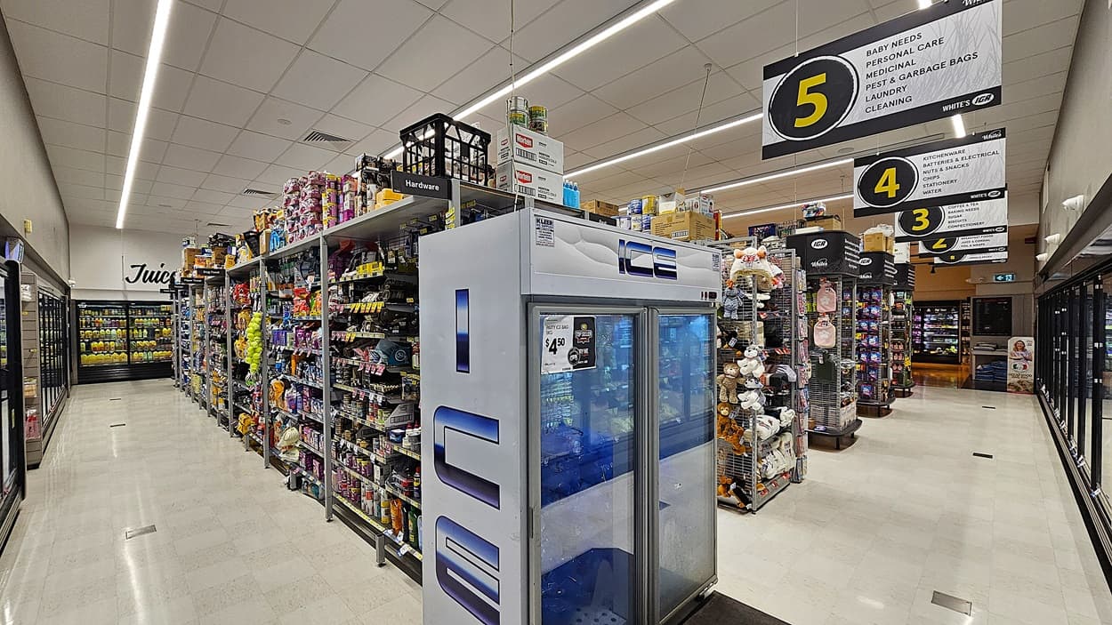 A supermarket aisle with a white tiled floor. On the left, there are refrigerated sections with glass doors displaying dairy products and juices. In the middle, there are shelves stocked with various household items, including light bulbs and batteries, and a large freezer labelled "ICE." The ceiling is white with rectangular fluorescent lights. On the right, there are more shelves with cleaning supplies and pet food, with overhead signs numbered 3 and 5 indicating different sections.