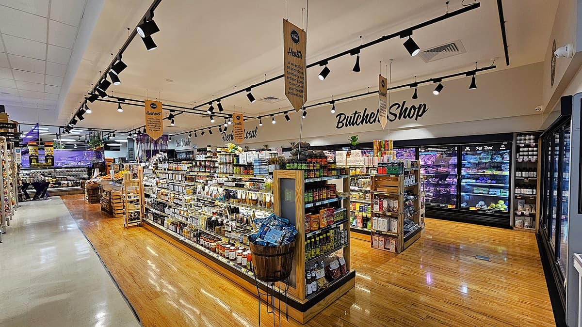 A supermarket with a mix of polished wooden and white tiled floors. On the left, there are shelves stocked with various packaged goods and a small wooden display table. In the middle, there is a large island with shelves filled with condiments, snacks, and a wooden barrel. The ceiling has white panels with black track lighting and hanging signs. On the right, there are refrigerated sections with glass doors displaying meats and dairy products, and a sign reading "Butcher Shop."