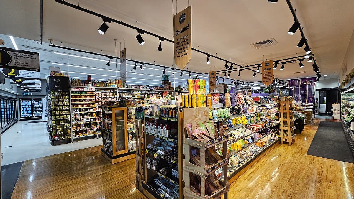 A supermarket with a mix of polished wooden and white tiled floors. On the left, there are shelves stocked with various packaged goods and a refrigerated section with glass doors displaying frozen items. In the middle, there are wooden display tables filled with snacks, condiments, and beverages, with hanging signs above. The ceiling has white panels with black track lighting. On the right, there are more shelves with fresh produce and a refrigerated section with dairy products.