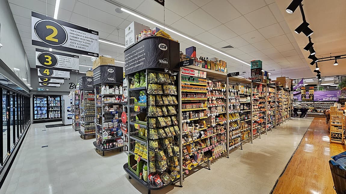 A supermarket aisle with a white tiled floor and a section of wooden flooring. On the left, there are tall shelves stocked with various snacks, including crisps and nuts, and a refrigerated section with glass doors displaying dairy products. In the middle, there are overhead signs numbered 2, 3, and 4 indicating different sections. The ceiling is white with rectangular fluorescent lights and black track lighting. On the right, there are more shelves filled with canned goods and condiments.