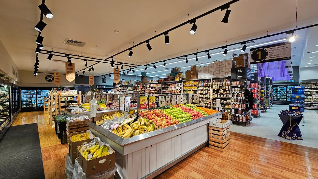 A supermarket with a mix of polished wooden and grey tiled floors. On the left, there are refrigerated sections with glass doors displaying beverages and dairy products. In the middle, there is a large display of fresh fruits, including bananas, apples, and oranges, with hanging signs above. The ceiling has white panels with black track lighting. On the right, there are shelves stocked with various packaged goods and a sign indicating aisle 1.