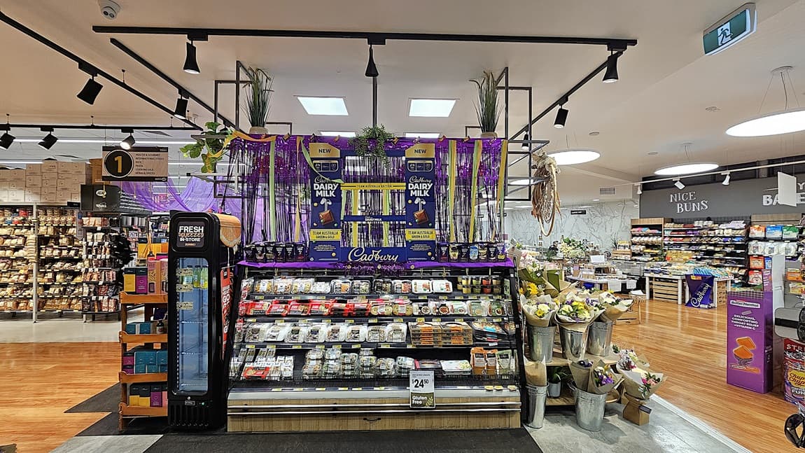 A supermarket with a mix of polished wooden and grey tiled floors. On the left, there is a black refrigerated unit with drinks and a sign indicating aisle 1. In the middle, there is a large display of Cadbury Dairy Milk chocolates, surrounded by purple decorations and hanging plants, with shelves of pre-packaged meals below. The ceiling has white panels with black track lighting. On the right, there are shelves with baked goods and a flower display in metal buckets.