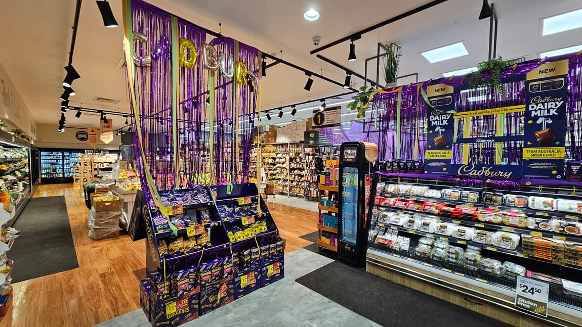 A supermarket with grey tiled and polished wooden floors. On the left, there is a large display of Cadbury Dairy Milk chocolates, decorated with purple streamers and hanging plants, with shelves of pre-packaged meals below. On the right, there are shelves with various food items, a fire extinguisher, and a sign reading "FOOD." The ceiling has white panels with black track lighting. In the background, there are more aisles, a flower display, and a sign that says "BAKE FOR MORE."