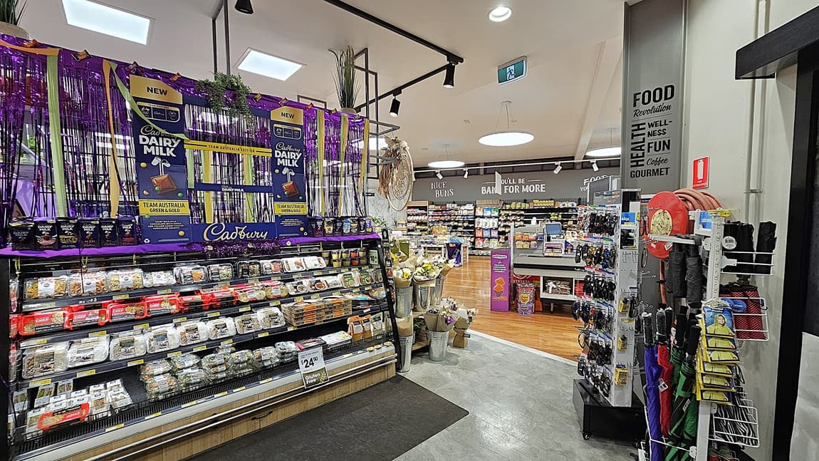 A supermarket with a mix of polished wooden and grey tiled floors. On the left, there are shelves with various packaged goods and a refrigerated section with glass doors displaying beverages. In the middle, there is a large display of Cadbury Dairy Milk chocolates, decorated with purple streamers and hanging plants. The ceiling has white panels with black track lighting. On the right, there are more Cadbury Dairy Milk products and a refrigerated section with pre-packaged meals.