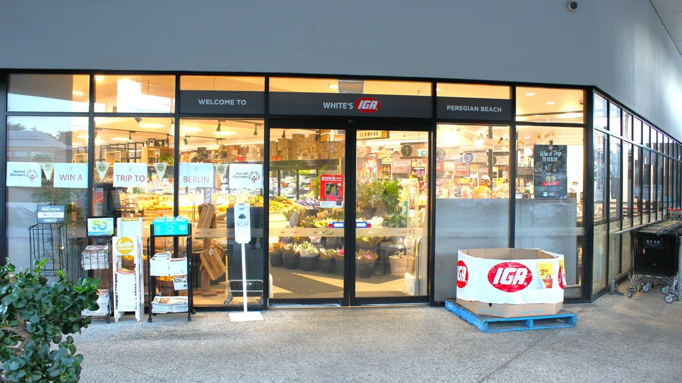 At the top of the automatic door of the shopping store is, “Welcome to White’s IGA Peregian Beach.” On the left side of the door is a thermal scanner with a hand sanitiser dispenser and a magazine rack. To the left of the store is a space for all the shopping carts.
