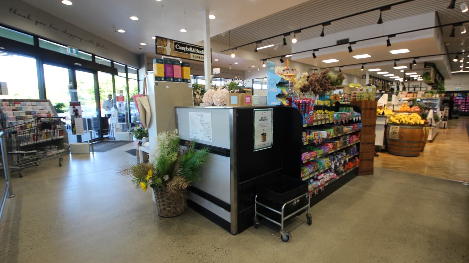 Closed checkout counter and a magazine rack near the main entrance of White’s IGA Peregian Beach. Near the counter is a white-painted wooden rectangular box with hooks for shopping bags hanging on the spaces in between. Right side of the image shows KitKat and Dairy Milk products. Next to this is the aisle for breakfast foods, coffee and tea, biscuits, jams and spreads, and health and wellness, as seen on the black circular signboard with the number 1.