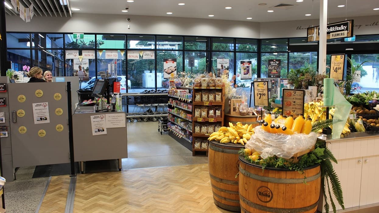 White’s IGA staff wearing black polos smiling beautifully at the assigned grey checkout counter near the main entrance of the store. In front of them is the chocolate and flower section, while to the right are bananas above brown barrels. Black shopping carts are also placed inside and near the checkout area. Seen through the glass walls are cars parked at the accessible parking spaces.