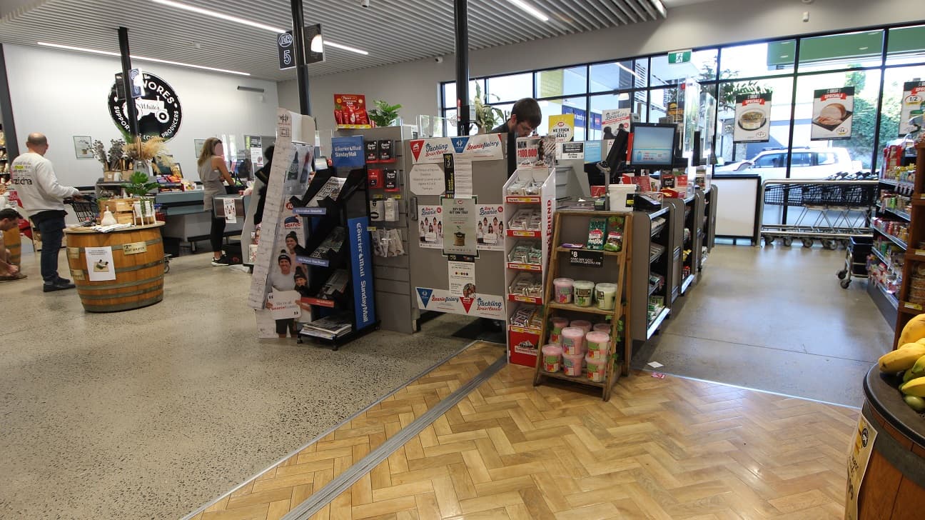 The image shows a long line at counter 6, while at Counter 5, there is a male staff. Compared to counter 6, counter 5 offers lots of other things to purchase, such as newspapers, beanies, candies, and chocolate bars. The ceiling in this specific section had thin, long, rectangular metal bars arranged parallel to each other and had spaces in between. A small part of the floor section is made of zigzagging blocks of wood with silver metal edges.