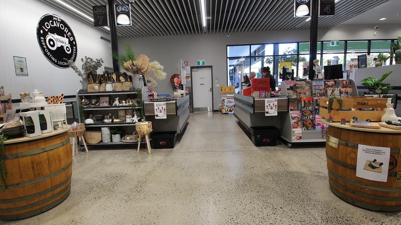 Side view of the closed checkout counter 7 and open counter 4 at White’s IGA Peregian Beach. There are two large wooden barrels directly placed opposite each counter. On counter 7, shoppers can also buy varying handcrafted designs and other home aesthetics, while on counter 4, there are several magazines to choose from on a grey wooden shelf. Above this shelf is the brand icon of Cérge with a QR code for customers with mobility disabilities.