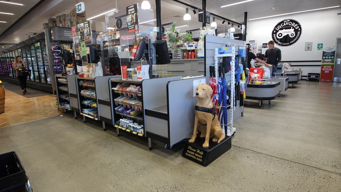 Exit pathway after payment on the checkout counters. On the left side is a brown dog statue, and behind it is a display of long and short umbrellas. Similar to other checkout counters, besides, there are some mints, chocolate, and candies on the grey shelves. There is a battery recycling unit placed against the white wall to the back. From this image, the granite floor covers the checkout counters while the zigzagging blocks of wood cover the series of refrigerators.
