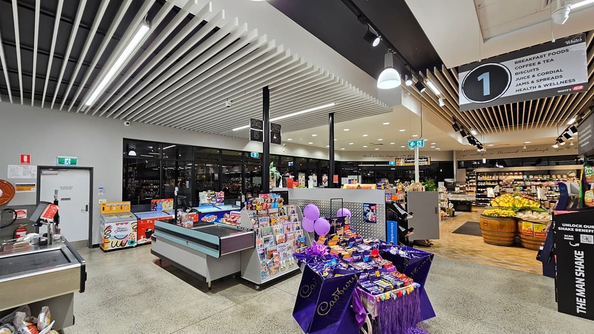On the left, there is a checkout counter with a display of magazines and snacks. In the middle, there is a table covered with a purple cloth displaying various confectionery items and balloons. In the background, there are shelves with packaged goods and a refrigerated section. On the right, there are wooden barrels filled with fruits and vegetables, including bananas and apples. The floor is polished concrete, and the ceiling has white slats with recessed and track lighting.
