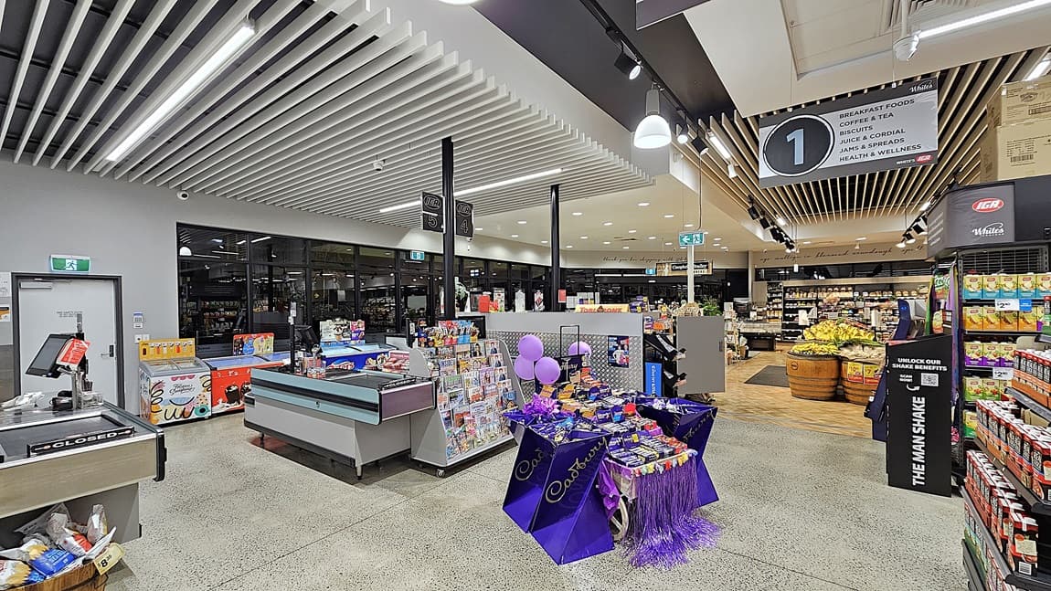 On the left is a checkout counter with a display of magazines and snacks. In the middle, there is a table covered with a purple cloth displaying various confectionery items and balloons. In the background, there are shelves with packaged goods and a refrigerated section. On the right, there are wooden barrels filled with fruits and vegetables, including bananas and apples, and more shelves with products. The floor is polished concrete, and the ceiling has white slats with recessed lighting