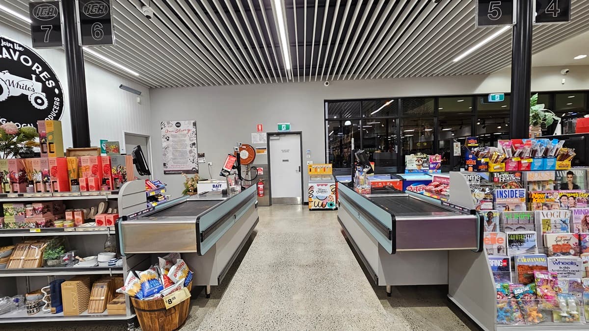 On the left, there is a checkout counter with a basket of snacks, a display of small items, and a register. In the middle, there are two checkout lanes with conveyor belts and a door in the background. On the right, there is another checkout counter with a large display of magazines and confectionery items. The floor is polished concrete, and the ceiling has white slats with recessed lighting.