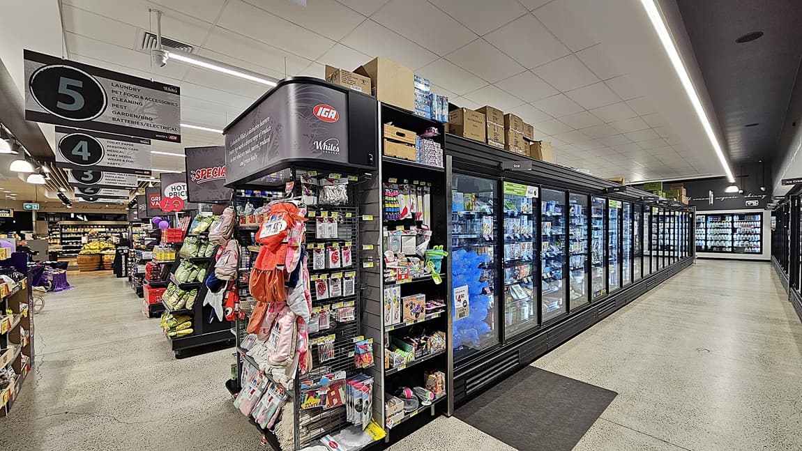 On the left, there are shelves with various packaged goods, including snacks and household items, and signs for aisles 3, 4, and 5. In the middle, there is a black shelving unit with pet supplies and a refrigerated section with glass doors containing frozen foods. In the background, there are more shelves and a checkout area. On the right, there are additional refrigerated display cases with glass doors. The floor is polished concrete, and the ceiling is white with linear fluorescent lighting.