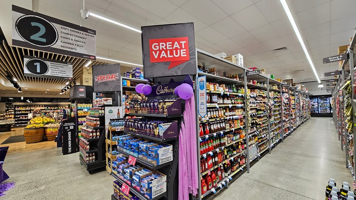 On the left, there are shelves with various snacks and a sign for aisle 2, which includes soft drinks, confectionery, crisps, and desserts. In the middle, there is a display with a "Great Value" sign and purple decorations, featuring Cadbury chocolates. In the background, there are wooden produce bins filled with fruits and vegetables. On the right, there are long shelves stocked with sauces, condiments, and other packaged goods. The floor is concrete, and the ceiling has fluorescent lighting.