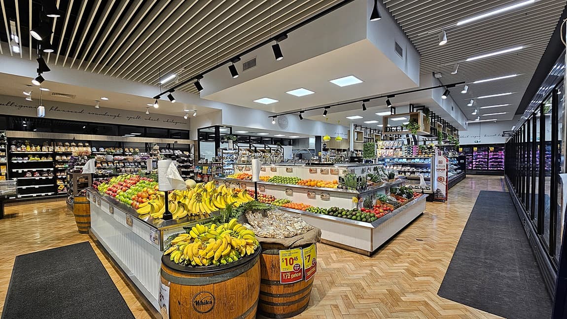On the left, there are wooden barrels filled with bananas and a produce section with various fruits and vegetables. In the middle, there are more produce displays, including tomatoes, peppers, and cucumbers, with a white counter and overhead lighting. In the background, there are shelves with packaged goods and a deli counter. On the right, there are long refrigerated display cases with glass doors. The floor is herringbone-patterned wood, and the ceiling has white slats with recessed lighting.