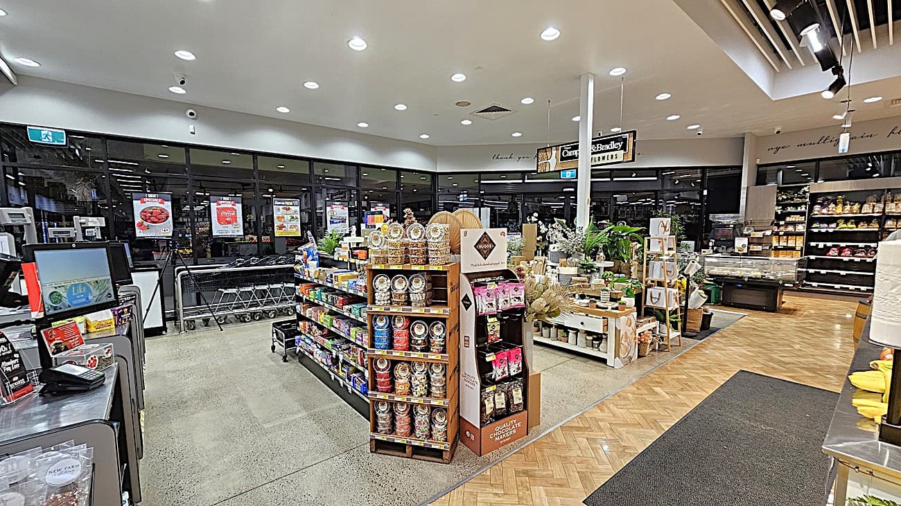 On the left, there are checkout counters with a display of magazines and snacks. In the middle, there are shelves with various packaged goods, including jars and boxes, and a small display of plants. In the background, there are large windows with shopping trolleys lined up against them. On the right, there are more shelves with packaged goods and a refrigerated section. The floor has polished concrete and herringbone-patterned wood, and the ceiling has recessed lighting with exposed beams.