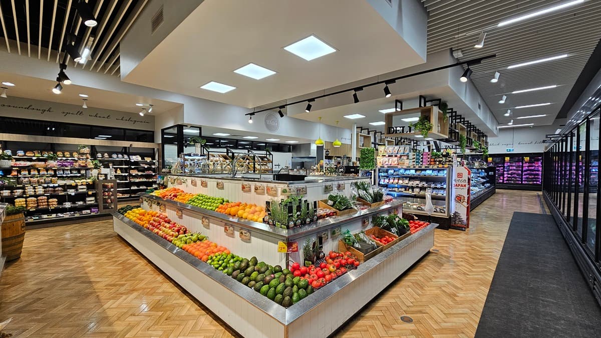 On the left, there are shelves with bread and baked goods. In the middle, there is a large produce display with a variety of fruits and vegetables, including avocados, tomatoes, and peppers, under square ceiling lights. In the background, there are more shelves with packaged goods and a section with dairy products. On the right, there are refrigerated display cases with glass doors containing drinks and other chilled items. The floor is herringbone-patterned wood, and the ceiling has white slats