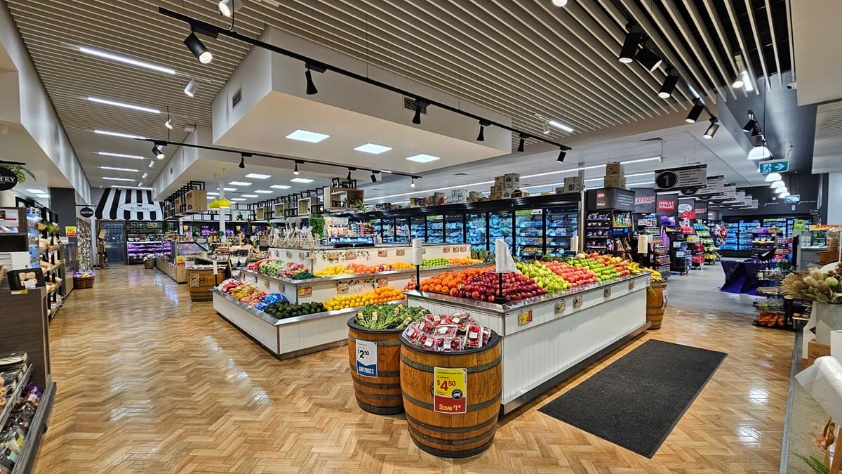 On the left, there are shelves with various packaged goods and a display of flowers. In the middle, there is a large produce section with wooden barrels filled with apples and a white counter displaying a variety of fruits and vegetables, including apples, oranges, and peppers. In the background, there are more shelves with products and a refrigerated section. On the right, there are additional refrigerated display cases with glass doors. The floor is herringbone-patterned wood.