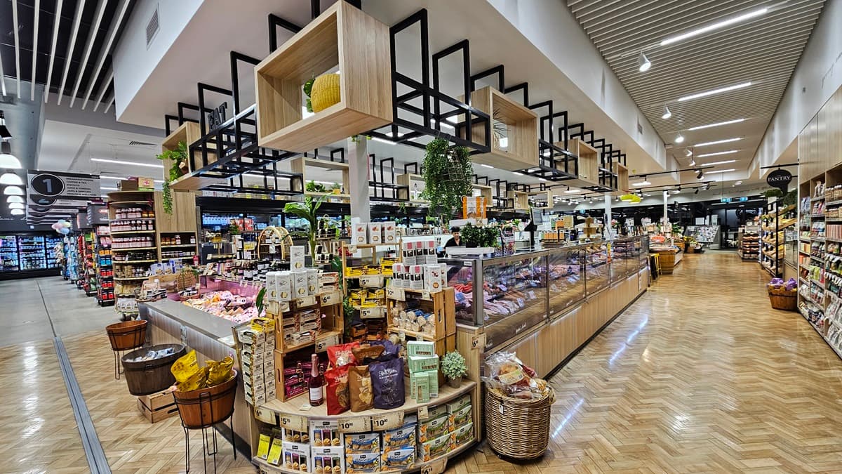 On the left, there are wooden barrels with packaged snacks and shelves with various goods, including jars and boxes. In the middle, there is a large display with wooden cubbies holding plants and decorative items above a counter filled with packaged foods and baked goods. In the background, there are more shelves and a refrigerated section. On the right, there are long refrigerated display cases with glass doors and additional shelves with products. The floor is herringbone-patterned wood.