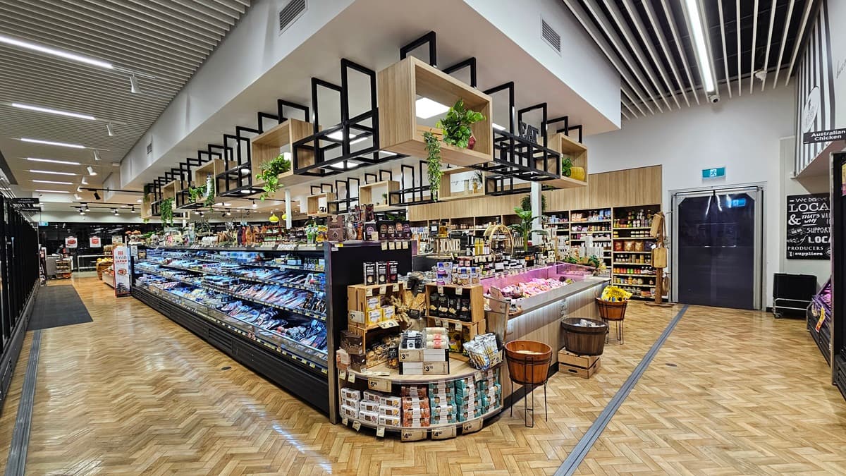 On the left, there are long refrigerated display cases with glass doors containing various chilled products. In the middle, there is a large island with fresh produce, including fruits and vegetables, and a display of packaged goods and baked items. Above the island, there are black-framed shelves with potted plants. In the background, there are more shelves with various products. On the right, there are additional shelves and a doorway. The floor is herringbone-patterned wood.