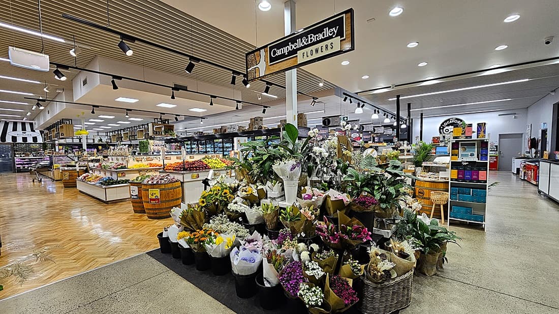 On the left, there are wooden produce displays with various fruits and vegetables, including apples and oranges. In the middle, there is a large flower display with bouquets and potted plants under a sign reading "Campbell & Bradley Flowers." In the background, there are more produce displays and shelves with packaged goods. On the right, there are additional shelves with products and a refrigerated section. The floor is a mix of polished concrete and herringbone-patterned wood.
