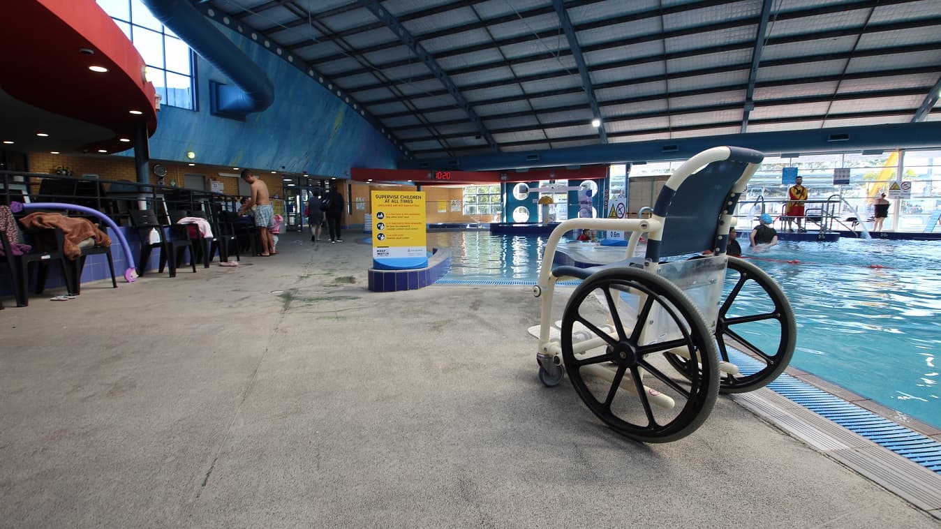 An aquatic wheelchair settled near the children’s pool. Black chairs are scattered around the pool for bags. At the end of the pools are glass walls that show the external pool area.