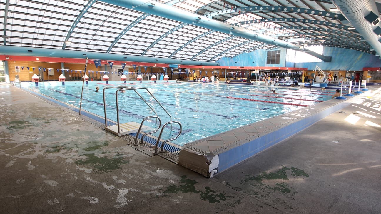 View of another end of the 8-lane 25m lap pool. The elevated section on the edge of the pool is made of blue tiles on the side, while on the top are diamond and triangular designs. Beside the pool ladder is a small white platform and metal handrail.