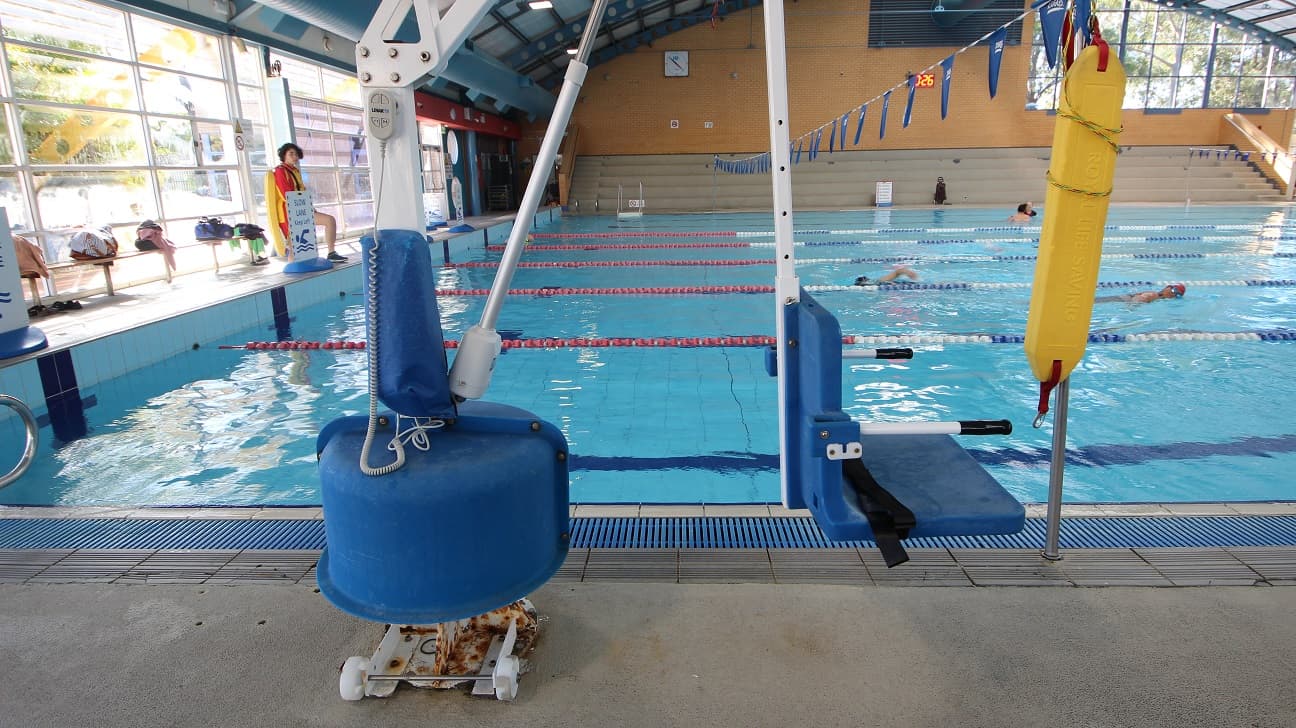 White pool hoist with blue seating chair at the 8-lane 25m lap pool. Near it is a yellow oblong floating platform used by lifeguards when saving someone. Long benches are placed against the glass walls for bags and other personal belongings.