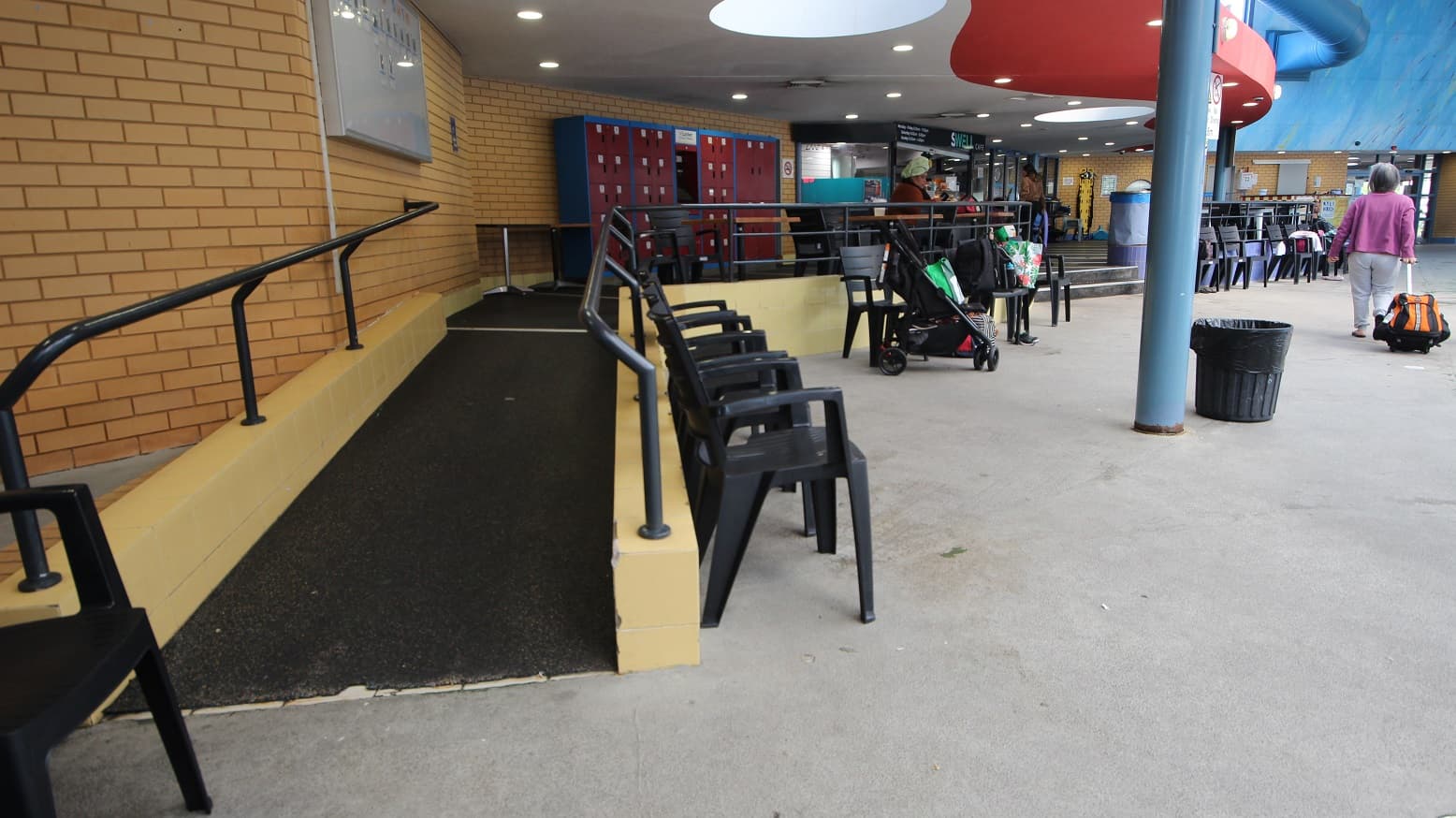 Entrance ramp with black smooth pathway, black handrails, and pale brown bricks leading to the red and blue locker cabinets and another seating area with mahogany table tops with metal frames and black chairs. A number of black chairs are designated slightly lower the ramp. The walls next to the Swell Café are made of bricks.