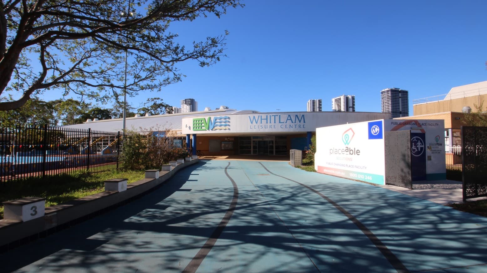A smooth blue concrete pathway leading to Whitlam Leisure Centre. The venue is a low-set building with grey, white, and blue painting. The upper wall of the front face of the building shows the name of the venue and its logo. On the left is a 50-meter external pool secured by a black fence, while on the right is a public changing place facility by Placeable Solutions. The entry point to the facility is through a black gate.
