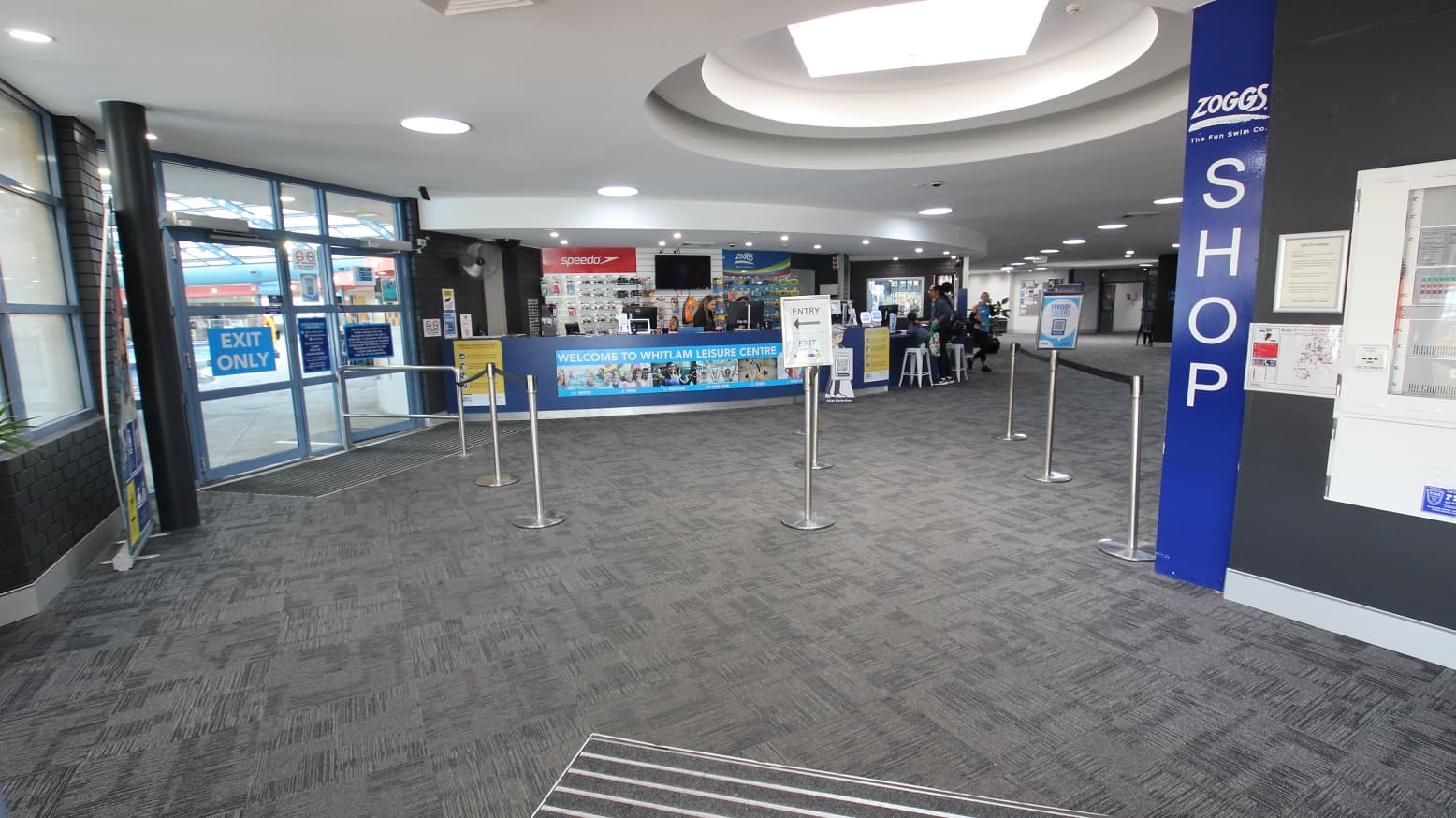 The image shows the view inside Whitlam Leisure Centre after passing through the entrance and exit doors which are separated by metal stanchions. There is a curved blue reception (with low counter accessibility) with a poster containing Welcome To Whitlam Leisure Centre. In front of the reception desk are stanchions and lane ropes for the queuing guests and members. This area has a grey brick wall, white ceiling, and grey, tiled floors with zigzag lines