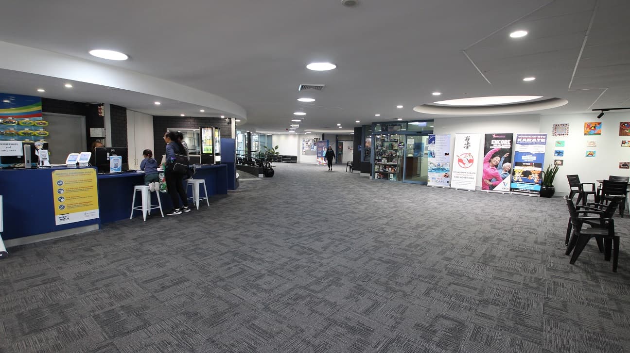 Next to the curvy reception tables is a thigh-light counter table with tall white bar stools. Heading towards the right from the reception desk are the Health Club and Swell Café. Different wall paintings and posters displayed near the wall can be seen opposite the reception area as well as black chairs and tables.