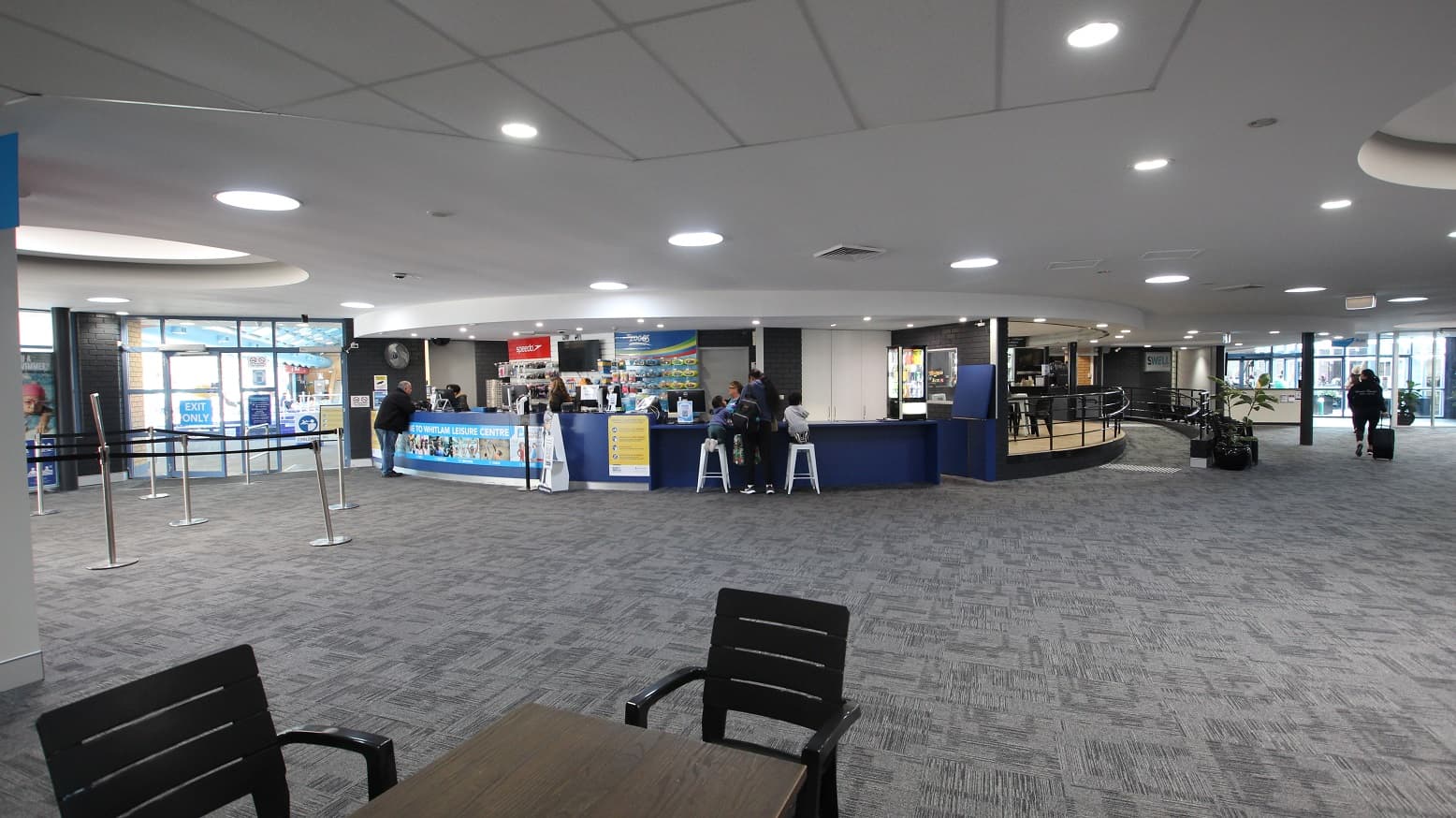 View when standing in the seating area containing black chairs and tables opposite of the reception desk. This image includes the peripheral part of the Swell Café with a low ramp-style entry point. The ramp has black metal handrails, and against it is a medium-sized potted plant and black chairs.