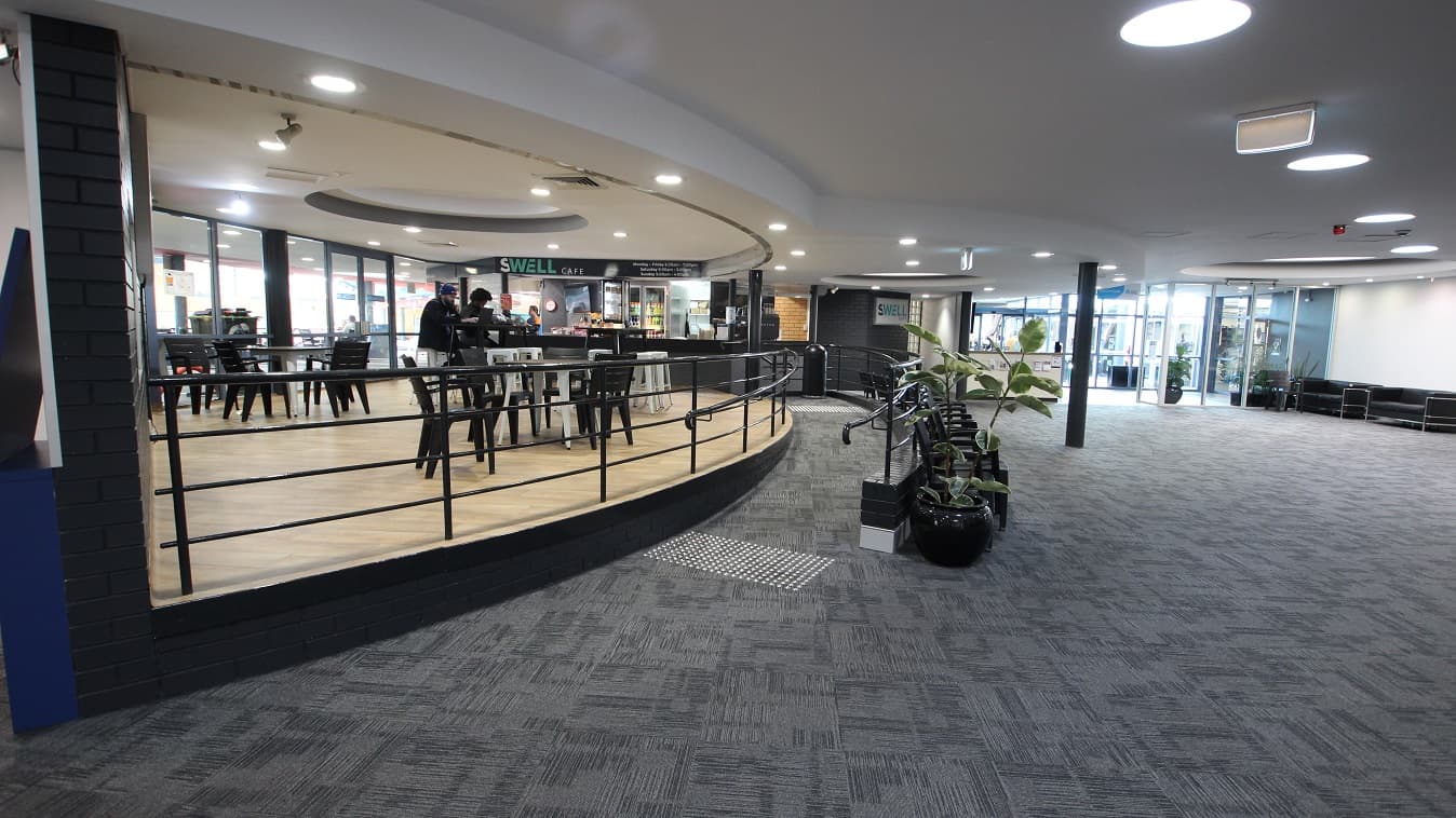 Low-ramp style entrance to the Swell Café. The café is a bit elevated from the ground floor and can be distinguished by its black and white tables & chairs and its walnut wooden floor. On the left side of the café are glass walls and doors which give a natural light coming from the outside.