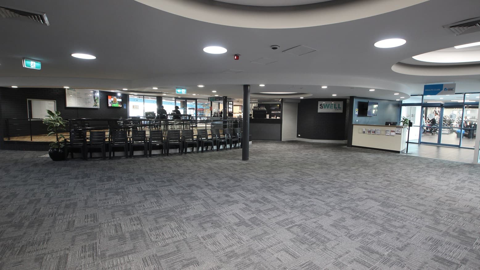 The image shows the Swell Café and Health Club. Just below the low-ramp style entrance of the cafe are several black chairs against the handrails. Heading straight after turning right from the reception desk is the Health Club, which can be identified by its white reception desk and blue wall. It has glass doors that allow guests to see other guests enjoying the Health Club facility.