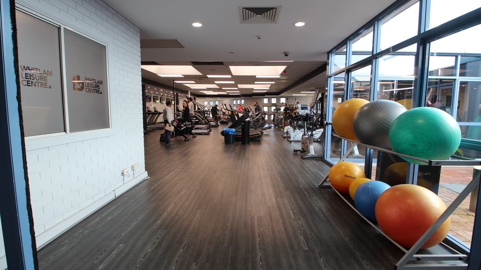 Left of the narrow corridor leading to the fitness facility is made of a white brick wall, while on the opposite side are glass walls and gym balls arranged in two layers of metal platforms. The first machines that guests could see were treadmills. The room is well-lit by square lights at the ceilings.