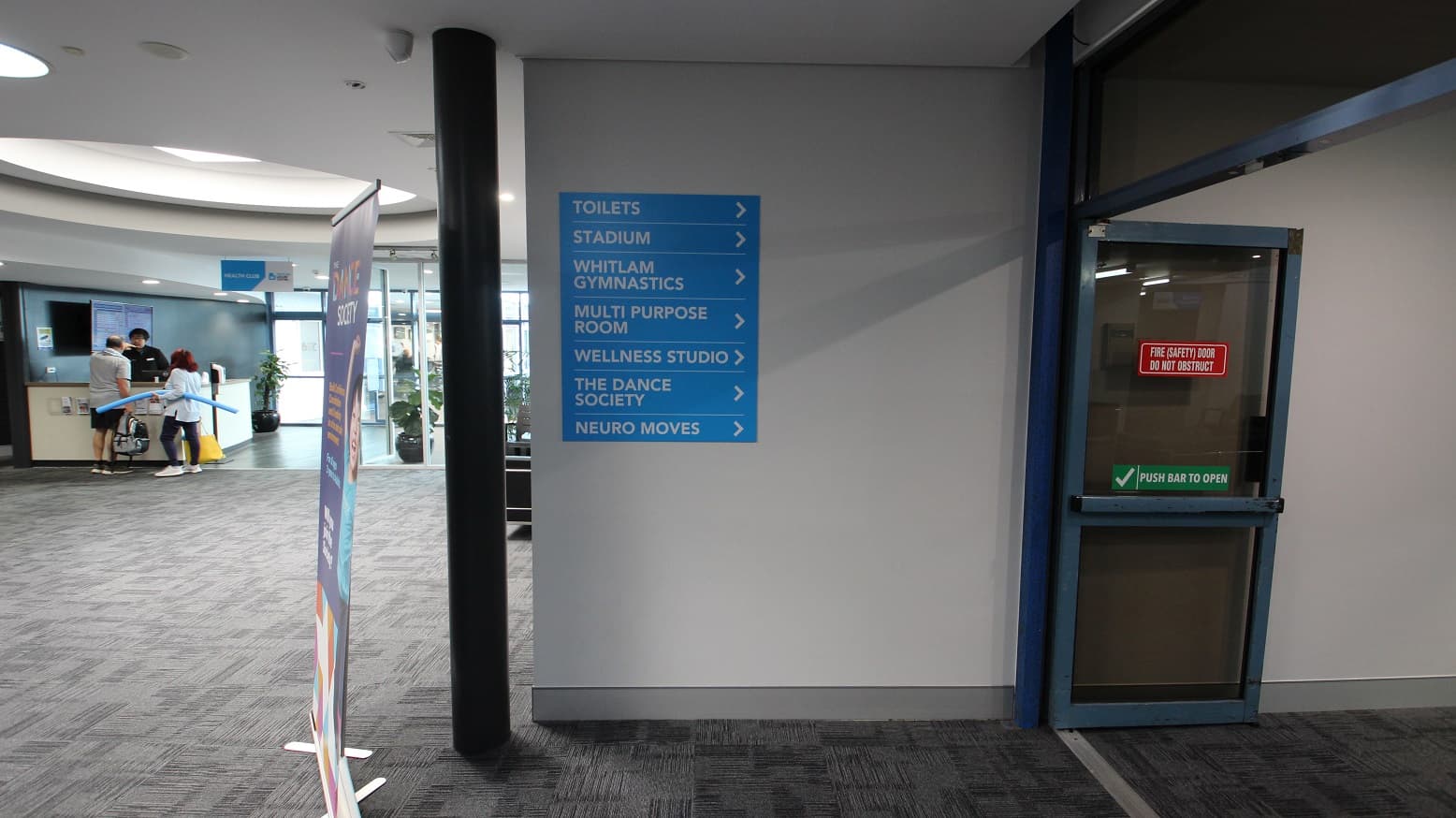Open, sliding, fire double doors. On the right of these doors is a wall with a class schedule for Bruins Liverpool Skills and Development basketball training and black chairs. There are also a number of chairs settled against the walls at the end of the corridor.