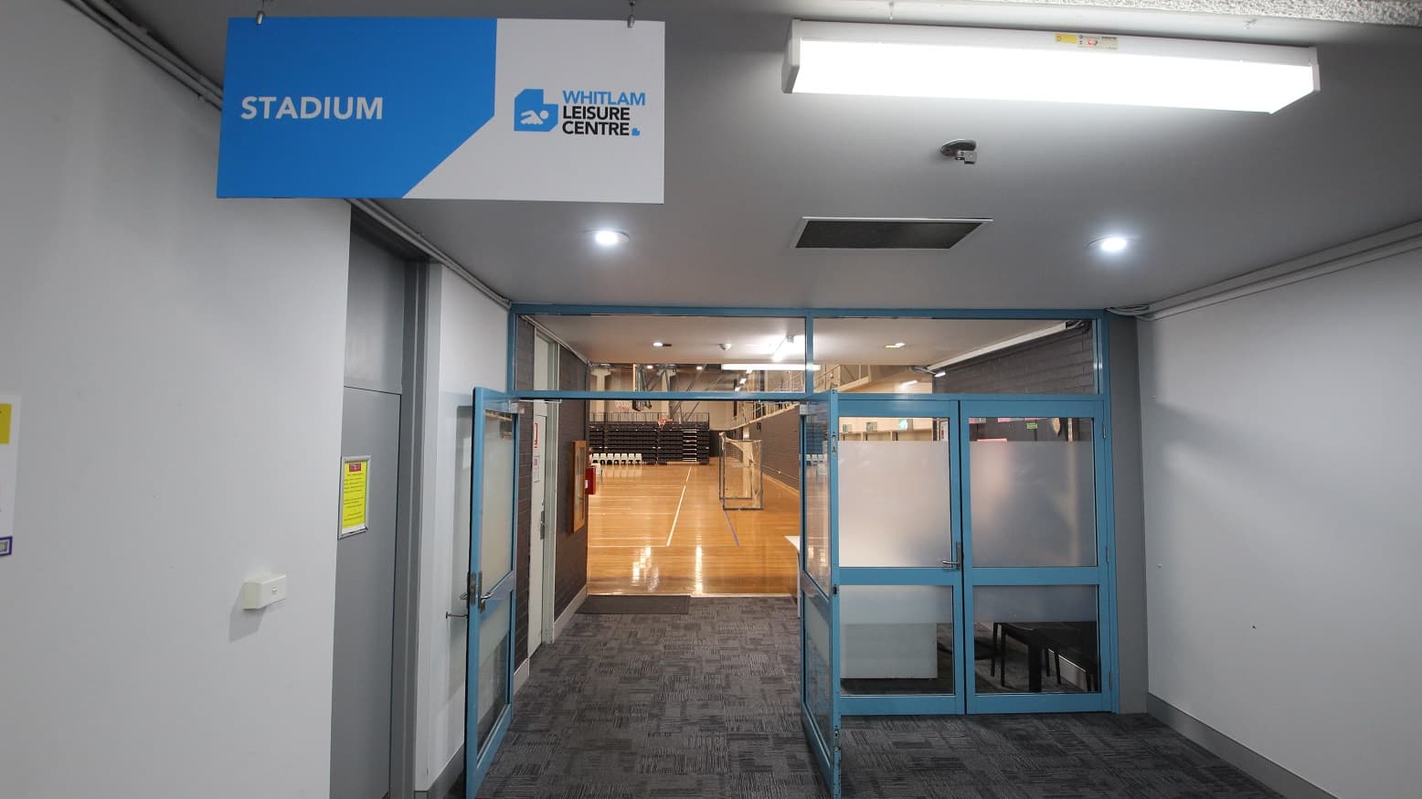 Entrance to the Stadium at Whitlam Leisure Centre. The double door on the left side is open, while the double door with a blurry portion on the right is not. Seen through the open door is the polished timber flooring, with a brown ceiling with dark beams, including fluorescent lig