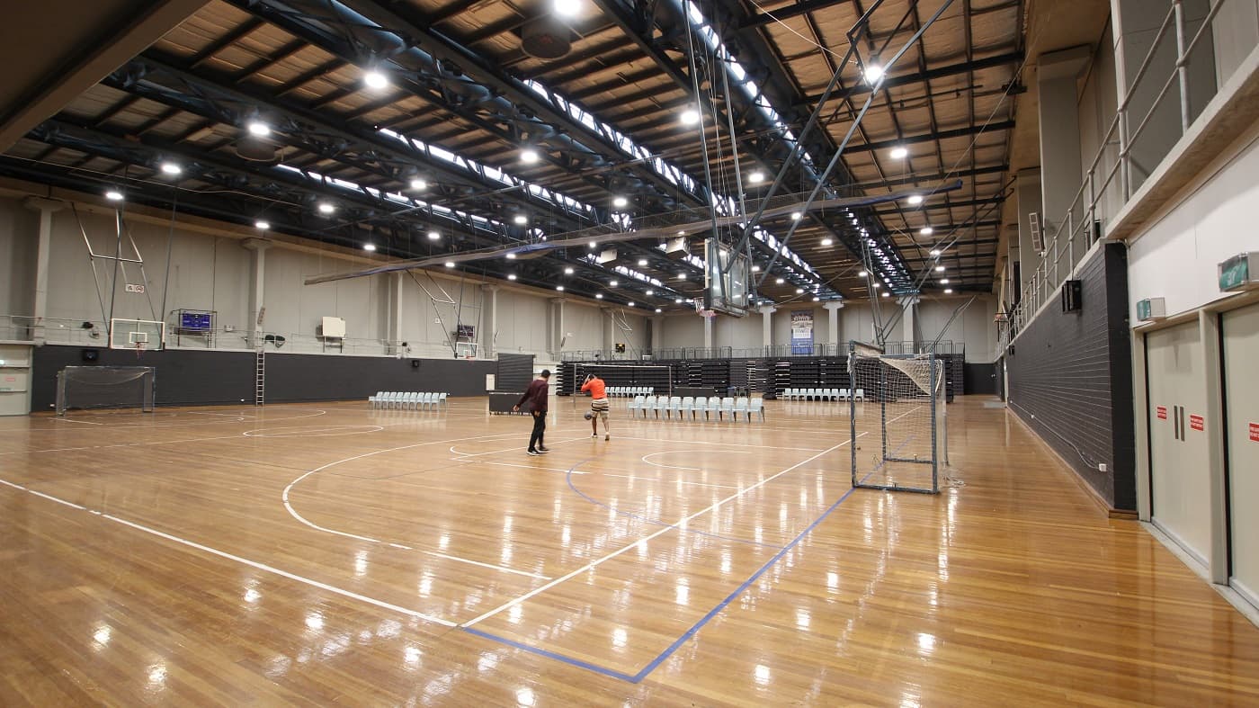 The Stadium is two basketball courts in size and has highly polished timber flooring, with a brown ceiling with dark beams including fluorescent lights. Underneath the basketball rings are football nets. Scattered on the other side of the basketball court are white chairs. The lower part of the walls are made of black bricks. The second floor with handrails can be accessed by a metal ladder.
