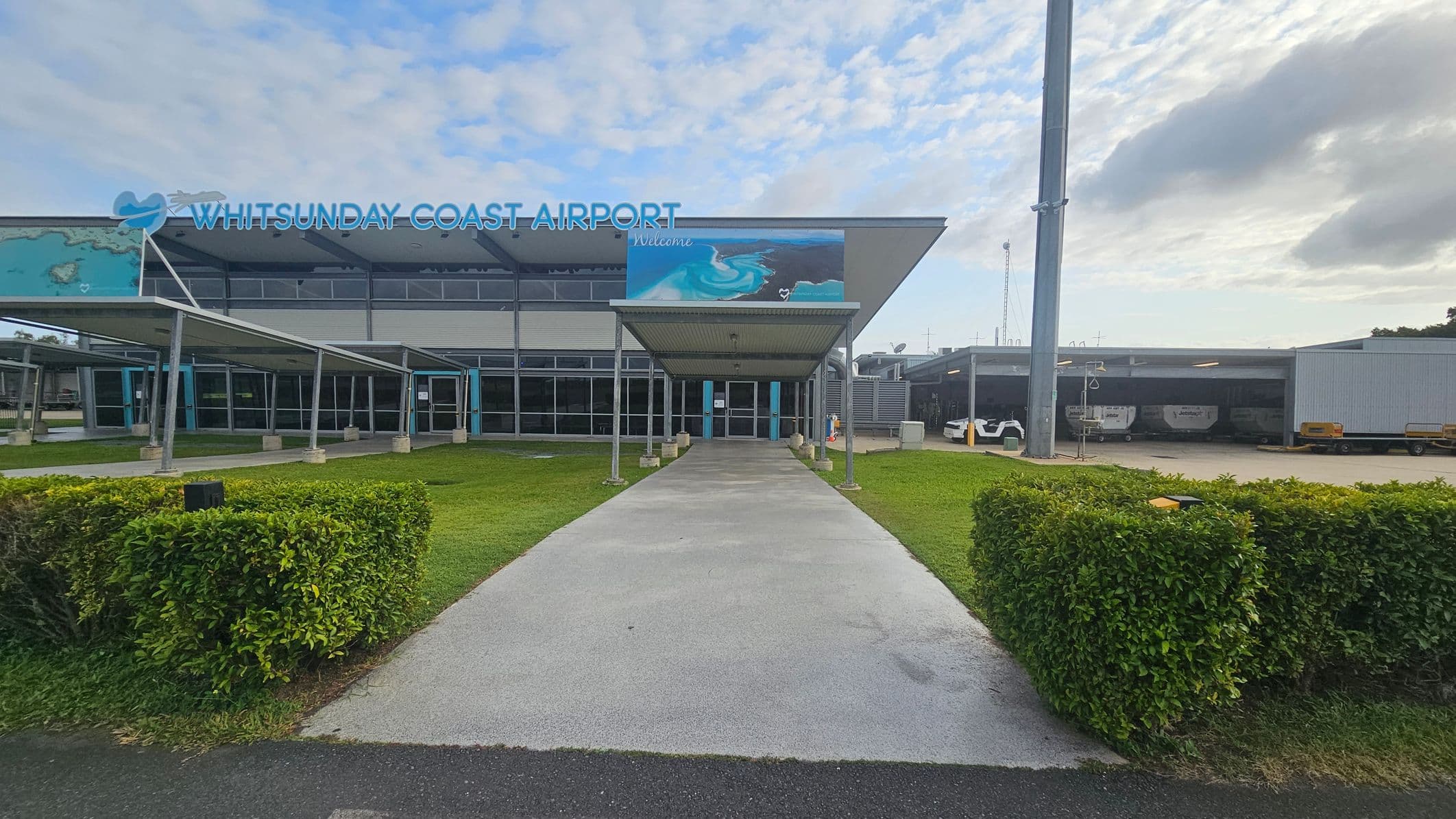 The image shows the entrance to Whitsunday Coast Airport. In the centre, a covered walkway leads to glass doors. The building has a large sign with the airport's name and a mural depicting ocean waves. On the left and right, there are green hedges and grass. To the right, a large pole and a covered area with vehicles are visible. The building has a modern design with a flat roof and large windows.