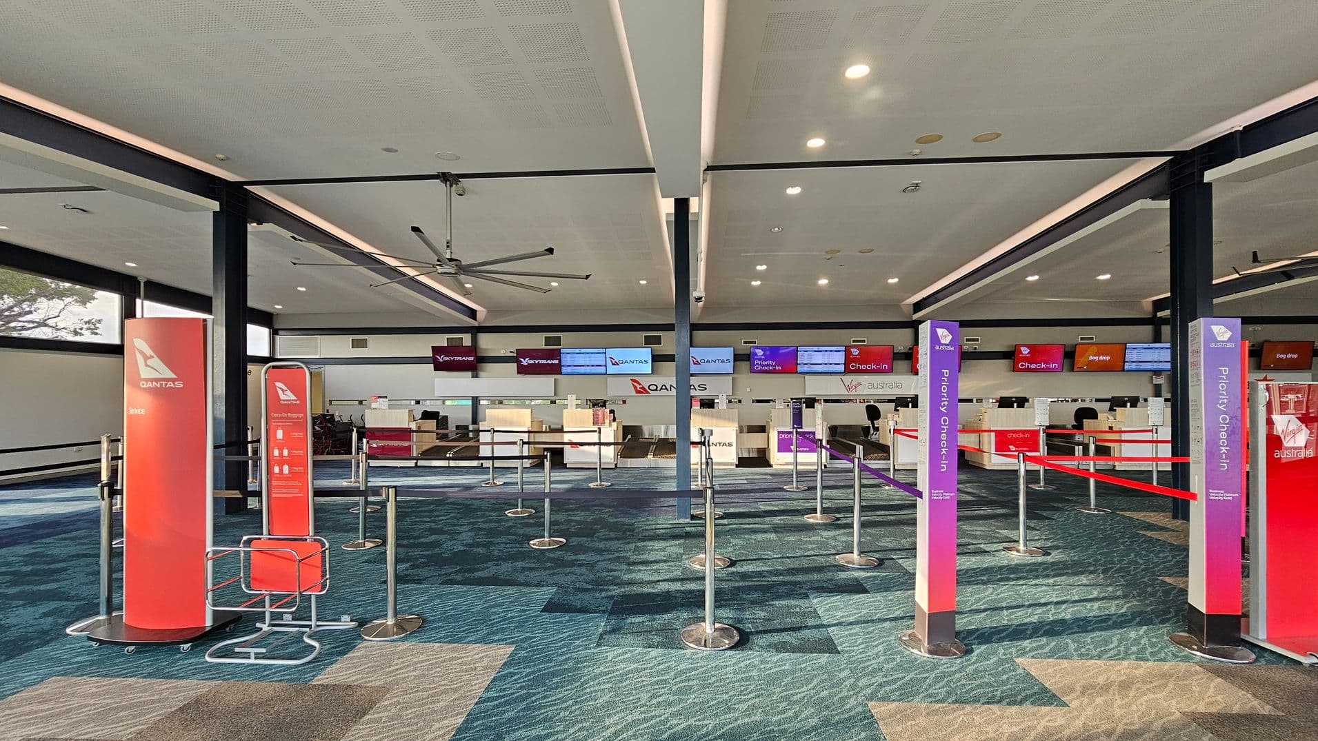 An airport check-in area with multiple counters in the background, featuring Qantas and Virgin Australia signage. The floor has a patterned carpet in shades of blue and beige. In the foreground, there are red and purple signs for Qantas and Virgin Australia, indicating service and priority check-in areas. The ceiling is light-coloured with recessed lighting and a large ceiling fan. Queue barriers are set up with retractable belts.